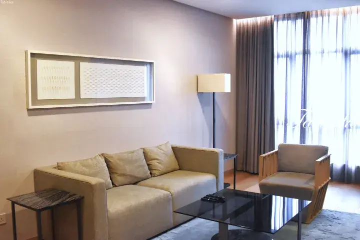 Modern hotel suite living area with beige sofa, glass coffee table, and large window with sheer curtains.