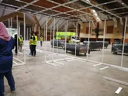 Workers in safety vests assemble white metal frames inside a large hall, with wrapped furniture and a green truck visible in the background.