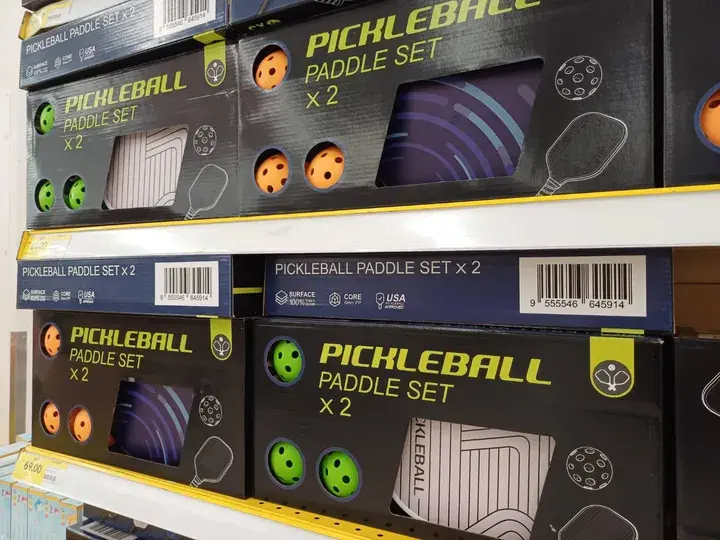 Boxes of Pickleball paddle sets displayed on retail shelves with visible balls and paddles.