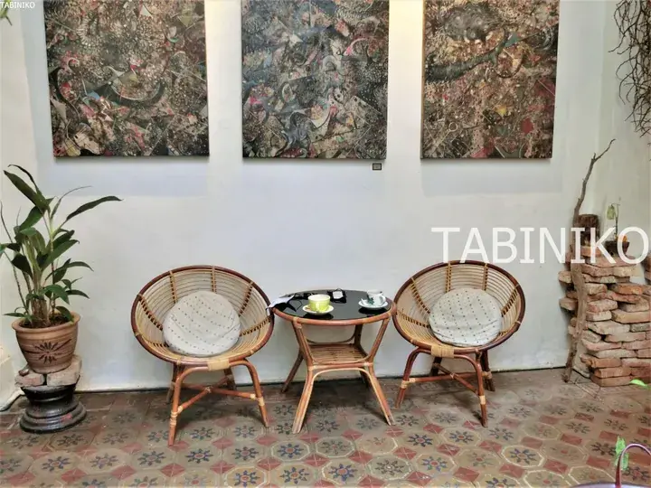 壁に抽象画が飾られたカフェのラタン製チェアとテーブル。TABINIKOのロゴが見えます。