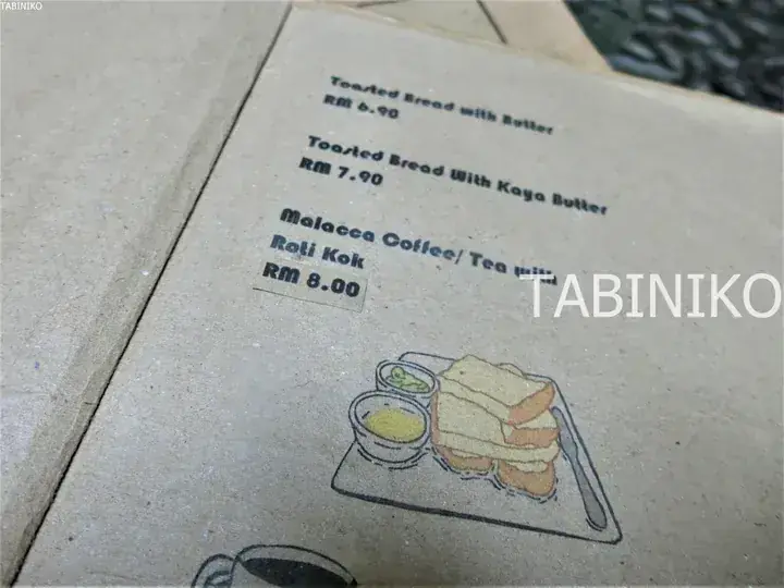 Menu card showing prices for Toasted Bread with Butter, Kaya Butter, and Malacca Coffee with Roti Kok at a Malaysian cafe.