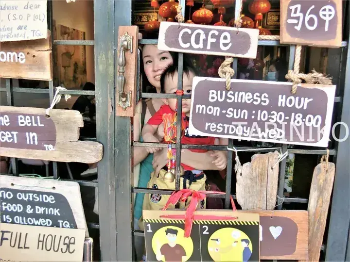 A woman and child peek through the barred window of a rustic cafe entrance adorned with wooden signs and red lanterns.