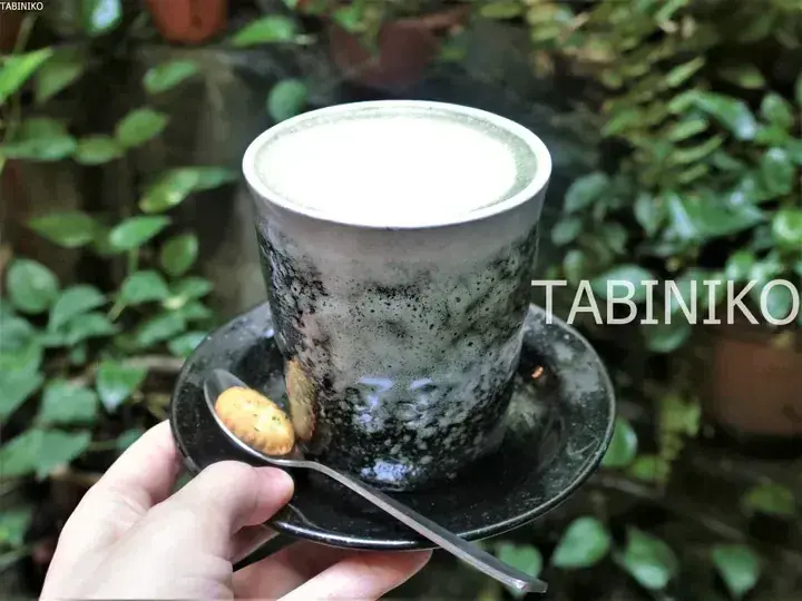 A hand holds a ceramic cup of frothy matcha latte with a biscuit on a saucer against a lush green garden backdrop.