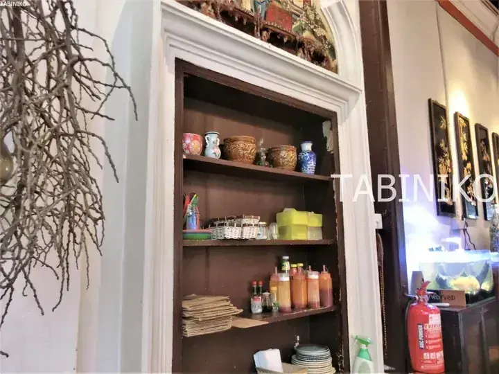 A built-in wooden shelf displays vintage ceramic jars, condiment bottles, and woven baskets in a cozy Malaysian cafe interior.