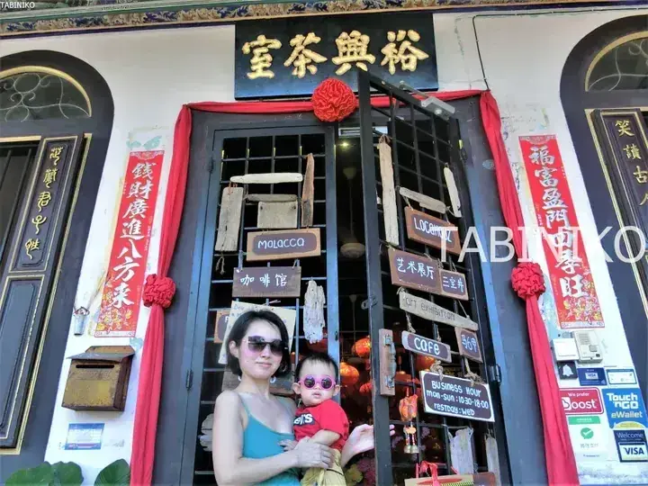 A woman holding a child wearing pink sunglasses stands in front of the Yu Xing Tea House entrance in Malacca, Malaysia.