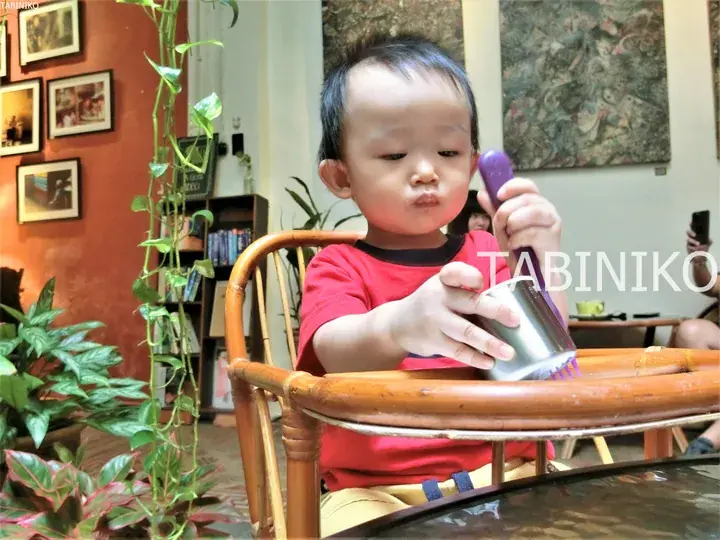 ラタン椅子に座り、金属製カップと紫色のブラシで遊ぶ赤いシャツの幼児。