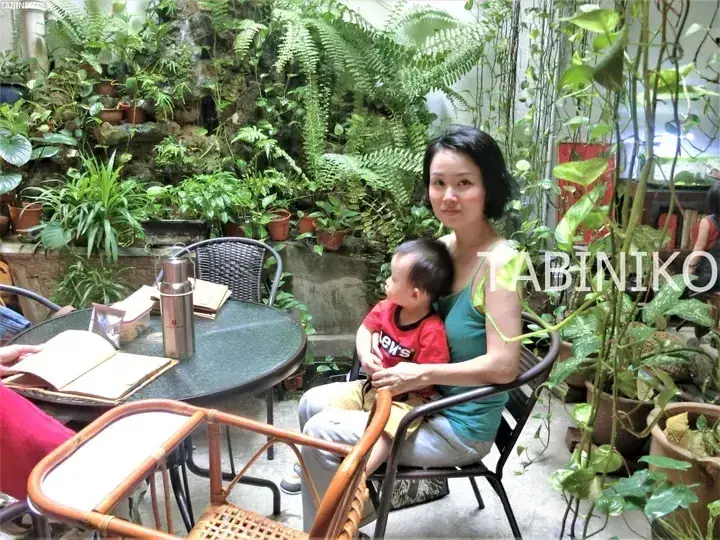 A woman and child sit at a table in a lush, plant-filled cafe with a rockery and wicker furniture.