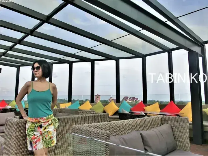Woman in sunglasses and floral shorts standing on a rooftop terrace with wicker furniture and colorful cushions overlooking a resort.