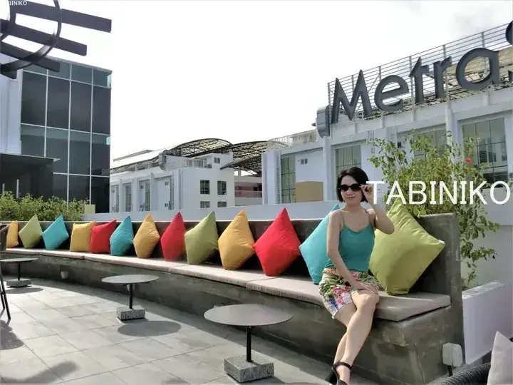 Woman sitting on a rooftop terrace bench with colorful cushions, with the Metrojaya building sign visible in the background.