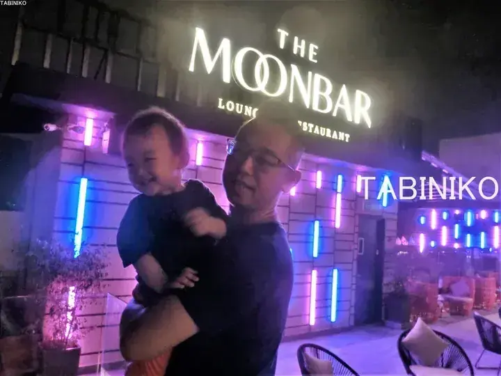 A man holding a smiling child in front of The Moonbar Lounge Restaurant with neon lights.