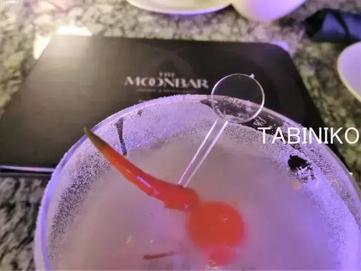 Frosted cocktail glass with red chili garnish and menu from The Moonbar in the background.
