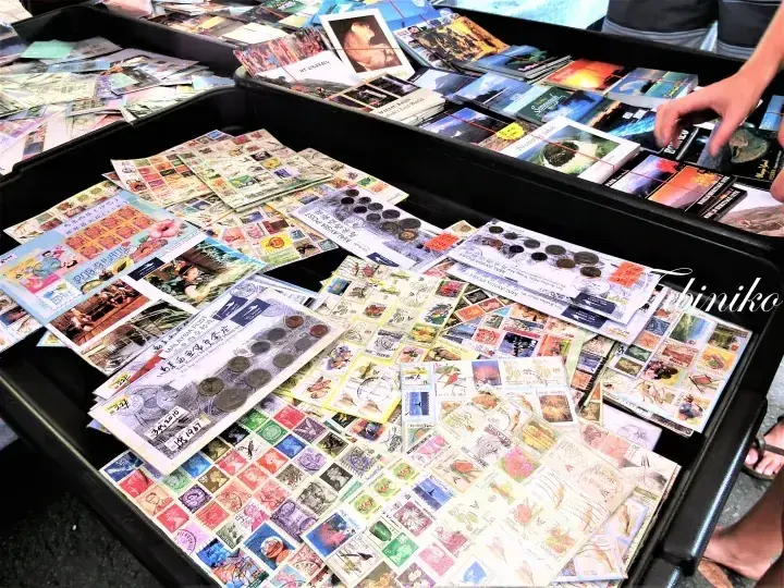 A market stall displays a large collection of Malaysian stamps and coins arranged on trays.