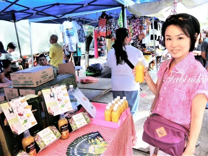 A woman at a market stall holds a bottle of Purigene juice, with boxes and brochures displayed on the table.