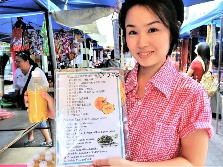 Woman holding a bottle of pumpkin juice and a sign listing its health benefits at a Malaysian market stall.