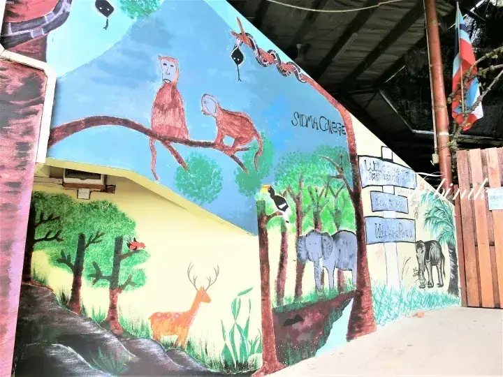 Vibrant wildlife mural at Sidma College featuring monkeys, elephants, and a deer with a sign pointing to the Amphitheatre and Wildlife Park.