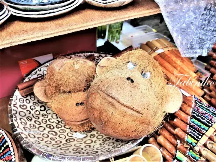 Handcrafted coconut shell monkey souvenirs displayed on a patterned tray at a Malaysian market stall.