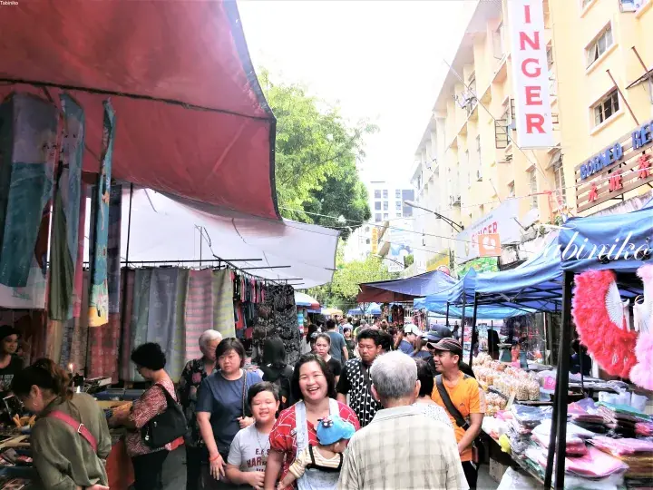 マレーシアの活気ある路上市場で赤と青のテントの下を買い物客が行き交う様子。
