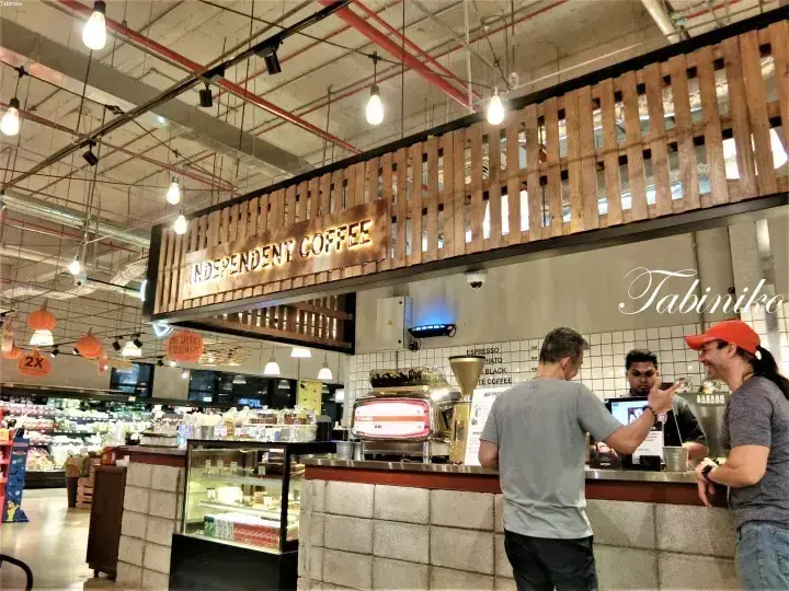 天井の配管が露出したモダンなスーパーマーケット内にある、木製アクセントのIndependent Coffeeカウンターで注文する客たち。