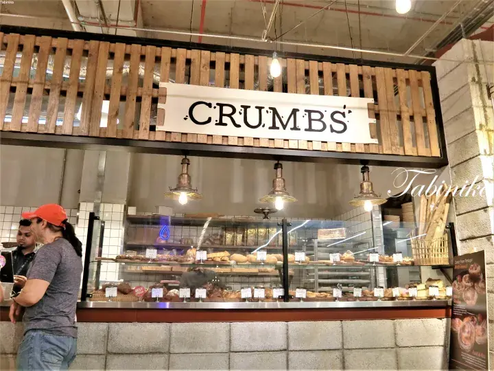 A bakery counter with a CRUMBS sign above a display case filled with pastries and customers waiting.