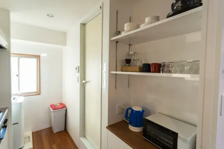 Modern kitchen pantry with open shelves holding coffee gear and a blue kettle, next to a washing area with a window.