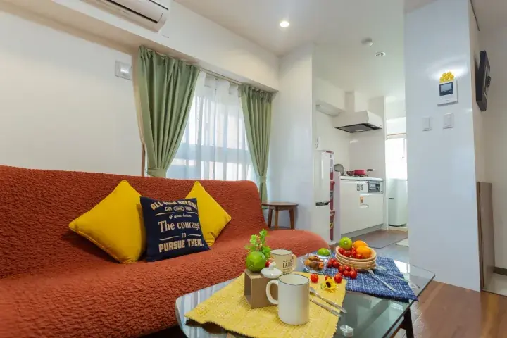 Cozy living room with orange sofa and yellow pillows, glass coffee table with fruit, and open-plan kitchen in the background.