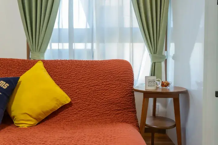 Cozy living room with an orange sofa, yellow cushion, and a wooden side table holding a mug near green curtains.