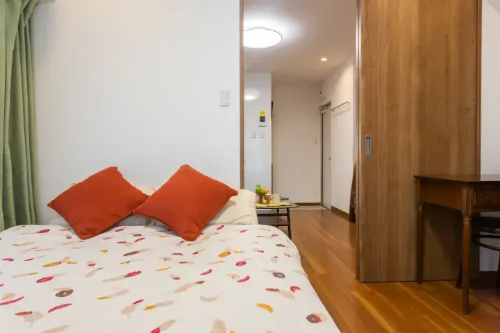 Cozy bedroom with patterned bedspread, orange pillows, and wooden flooring leading to a hallway.
