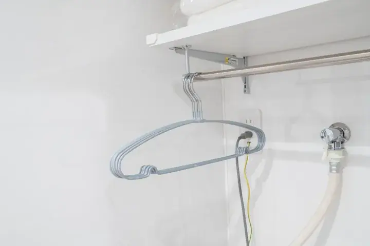 A grey wire hanger hangs on a metal rod in a white utility closet with a shelf and water pipe.