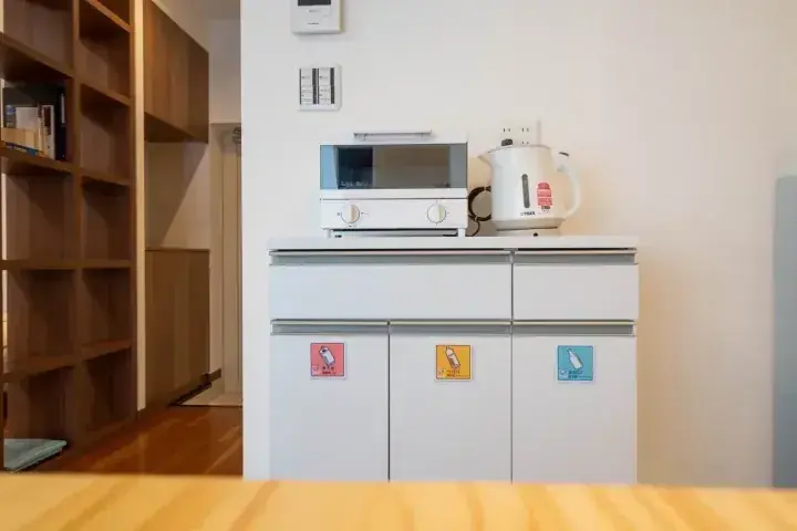電子レンジやか kettle が置かれた白いキッチンキャビネット。プラスチック、紙、ガラスのリサイクルステッカーが貼られている。