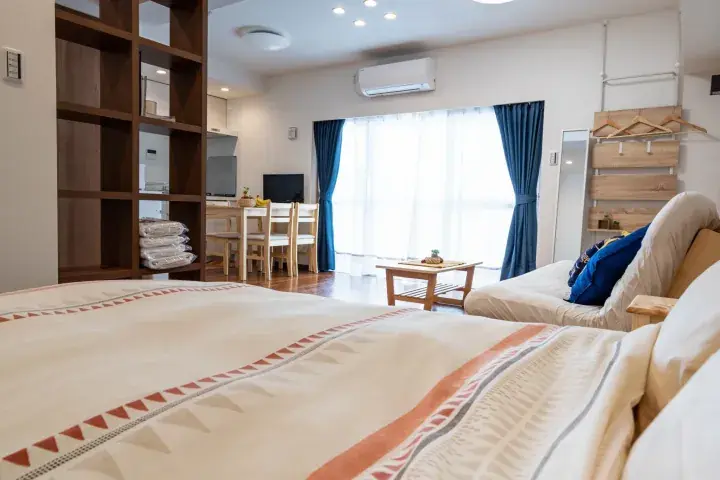 Bright studio apartment with a patterned bed, wooden shelving, dining area, and blue curtains.