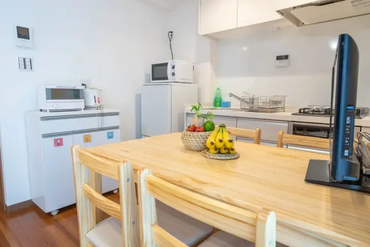 Modern kitchen with wooden dining table, bananas, fruit basket, and white appliances in a bright apartment.