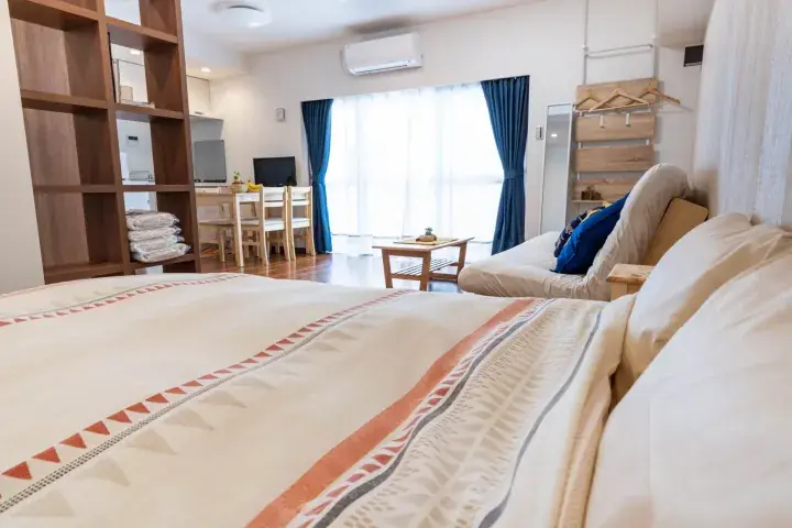 Bright modern studio apartment featuring a patterned bedspread, wooden shelving, dining area, and large window with blue curtains.