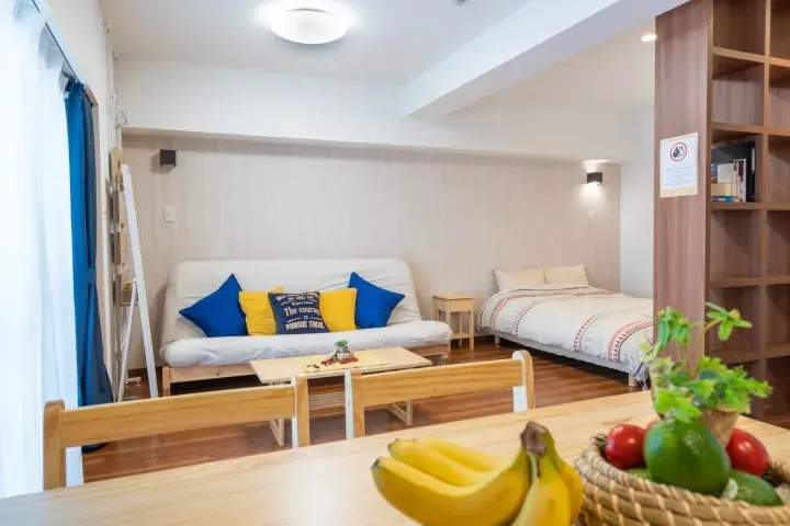 Bright studio apartment interior with a white sofa, wooden dining table, and fresh fruit on the counter.