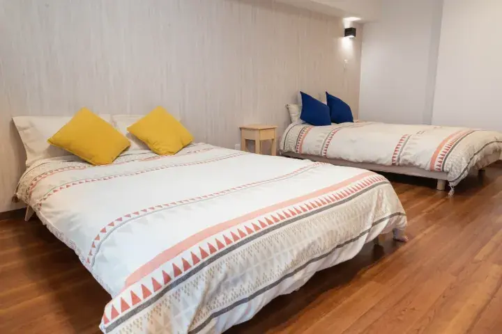 Bright bedroom with two beds featuring patterned duvets, yellow and blue pillows, and wooden flooring.