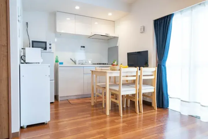Bright open-plan kitchen and dining area with wooden floors, white cabinetry, and a table set near a window with blue curtains
