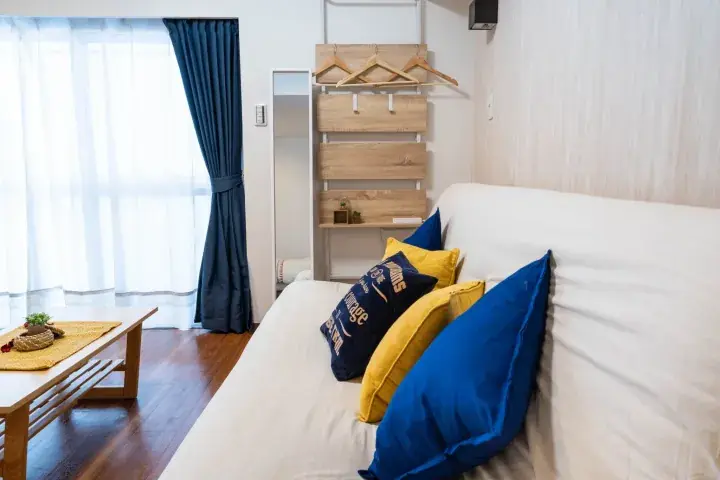 Bright living room with white sofa, blue and yellow cushions, wooden coffee table, and wall-mounted wooden shelf.
