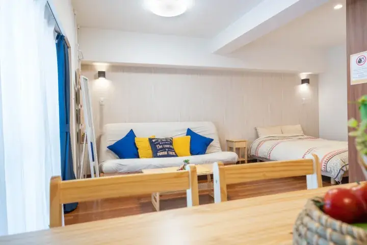 Bright studio apartment interior with a white sofa bed, blue and yellow cushions, and a single bed in a modern setting.