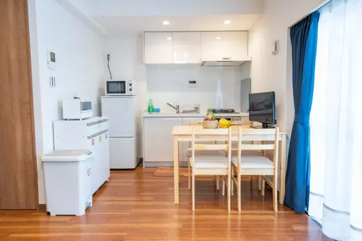 Bright studio apartment with white kitchen, wooden dining table, and blue curtains.
