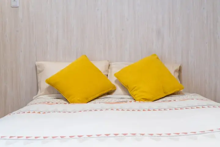 Neatly made bed with mustard yellow cushions and patterned duvet in a bright room with light wood paneling.
