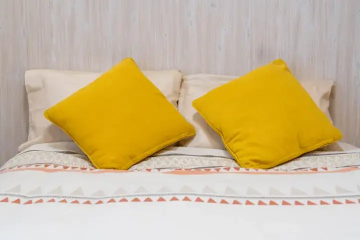Two mustard yellow cushions on a bed with patterned bedding against a light wood-paneled wall