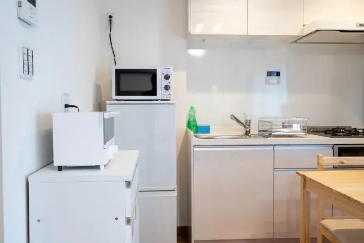 Bright white kitchen with microwave, refrigerator, sink, and wooden dining table in a modern apartment.