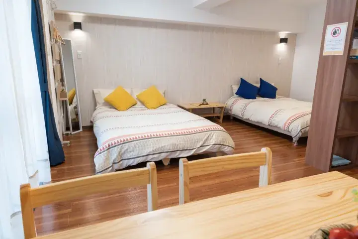 Bright bedroom with two beds, yellow and blue pillows, wooden furniture, and a dining table in the foreground.