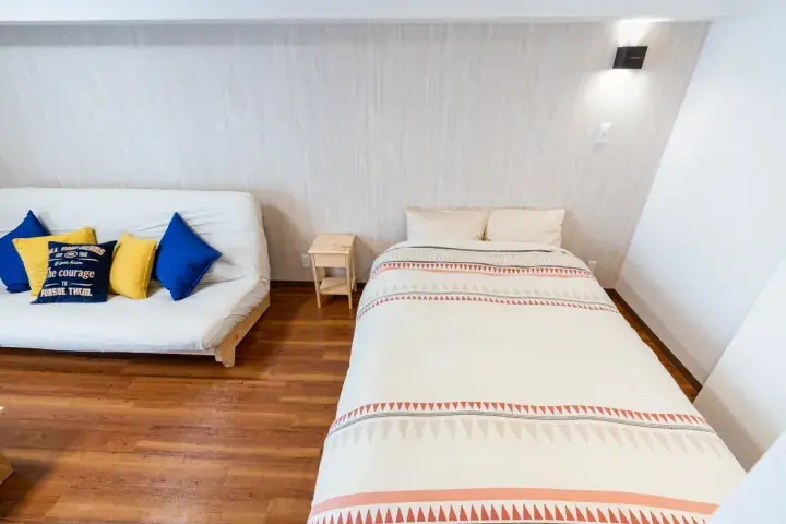 Bright bedroom with a patterned bedspread and a white sofa with blue and yellow cushions on wooden flooring