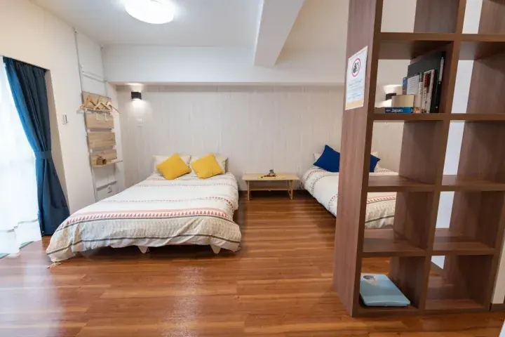 Bright bedroom with wooden floors, a double bed with yellow pillows, a single bed, and a wooden bookshelf divider.