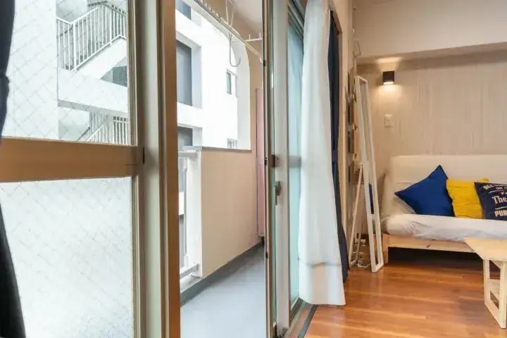 木製フローリングとソファのある明るいアパートの室内。バルコニーのドアが開き、白い建物が望める。
