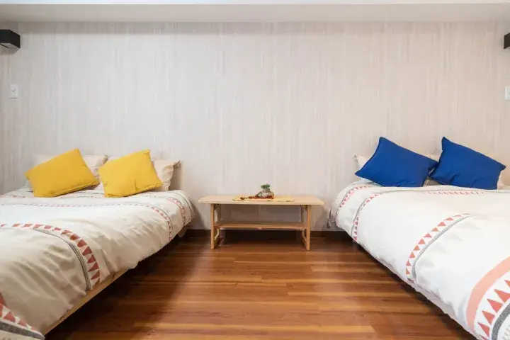 Bright bedroom with two single beds, yellow and blue pillows, and a wooden table on hardwood flooring.