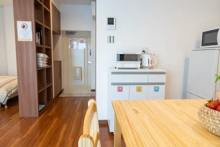 Modern apartment interior featuring a wooden dining table, kitchenette with microwave and kettle, and a hallway leading to the entrance.