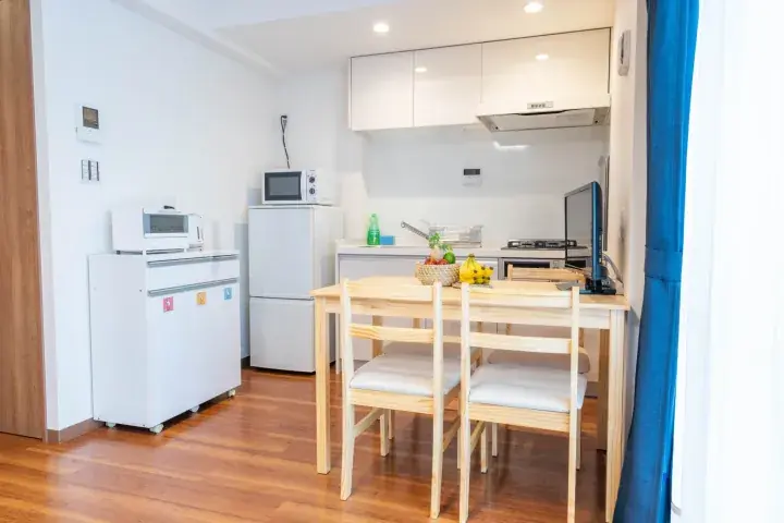 Bright studio apartment dining area with wooden table, chairs, kitchen counter, and white appliances.