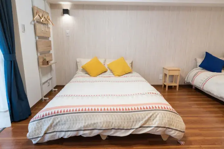 Bright bedroom with a double bed featuring yellow pillows and a patterned duvet, plus a single bed and wooden coat rack.