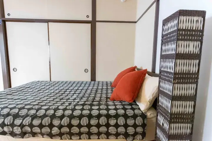 Bedroom with a patterned duvet, orange pillows, and a tall woven room divider in a traditional Japanese-style room.